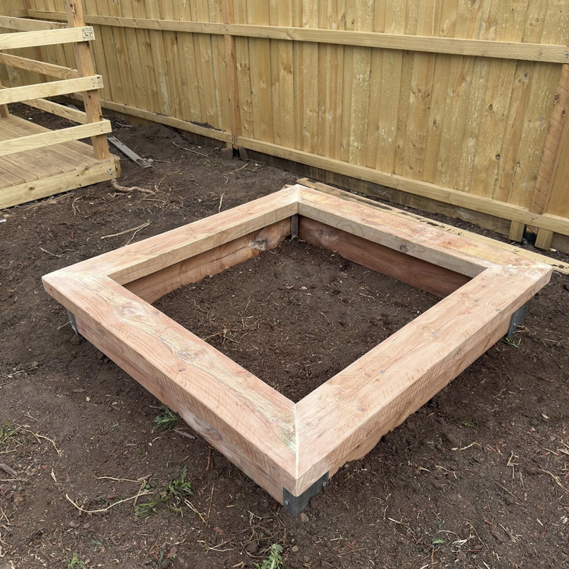 Square timber garden bed frame on bare soil beside a new paling fence