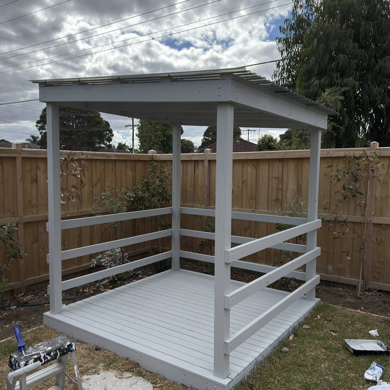 Small grey painted timber shelter with a roof, railings and deck floor in a backyard