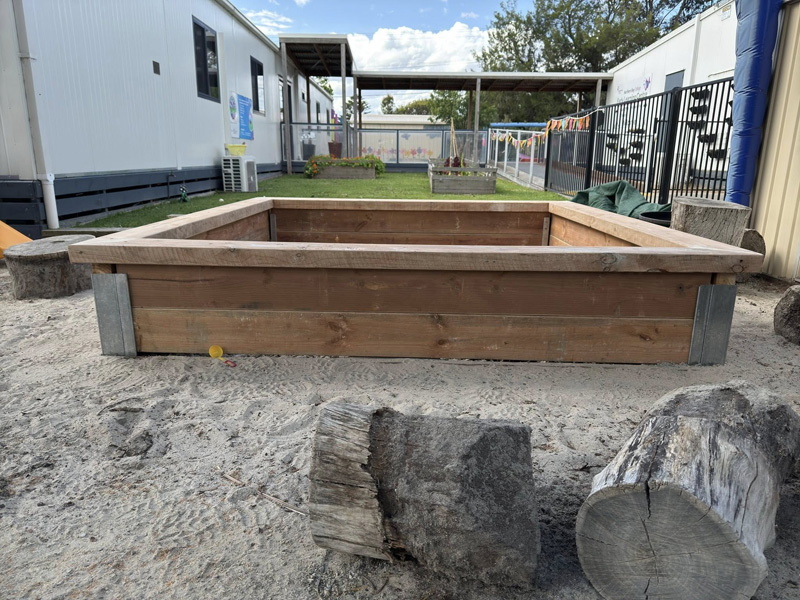 Large raised timber box frame in a sandy play yard with log stools