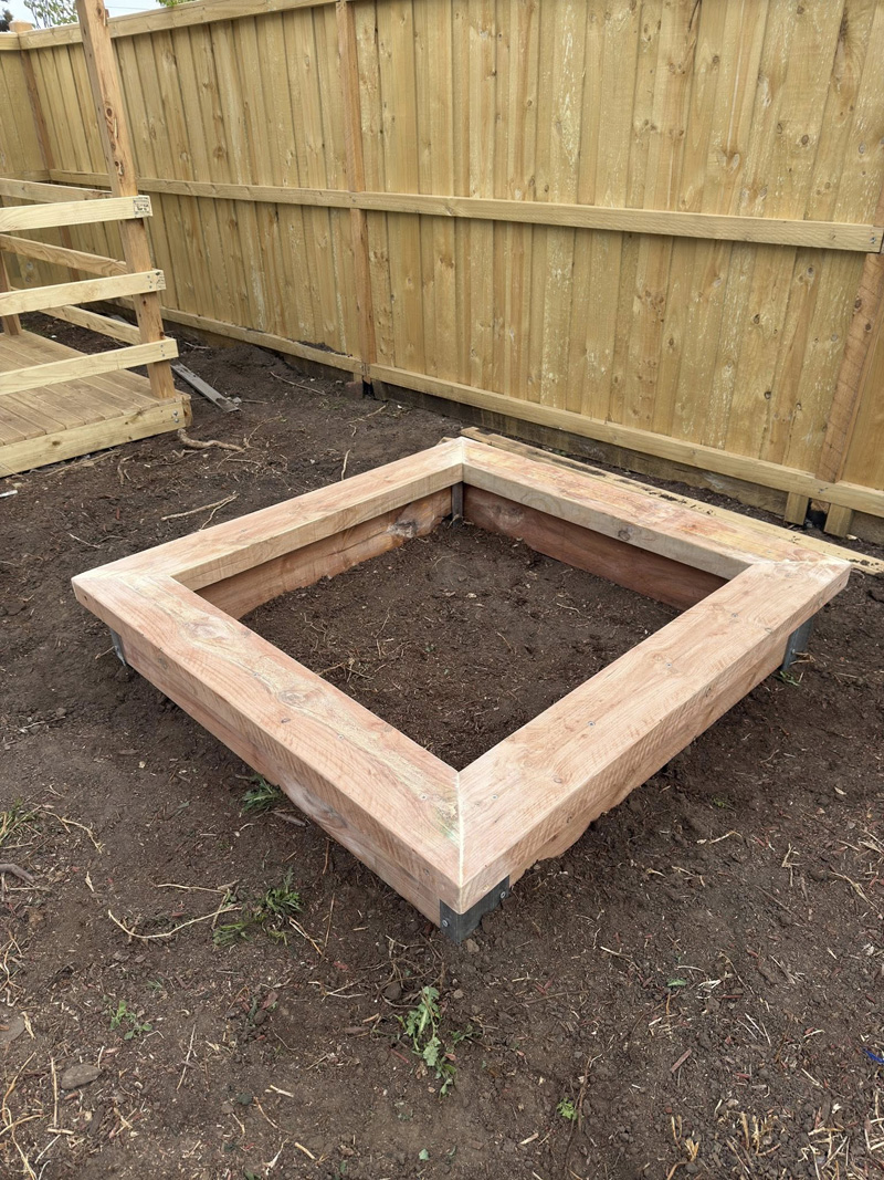 Square timber garden bed frame on bare soil beside a new paling fence
