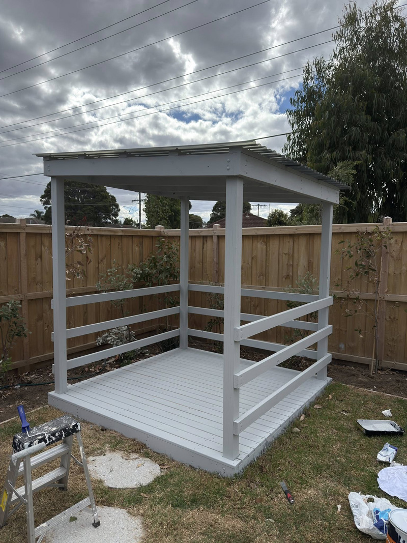 Small grey painted timber shelter with a roof, railings and deck floor in a backyard
